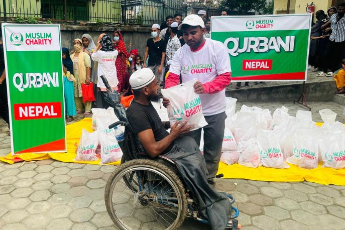 A volunteer hands a food bag labeled Muslim Charity to a person in a wheelchair. Banners reading Qurbani Nepal are displayed. Several people stand in