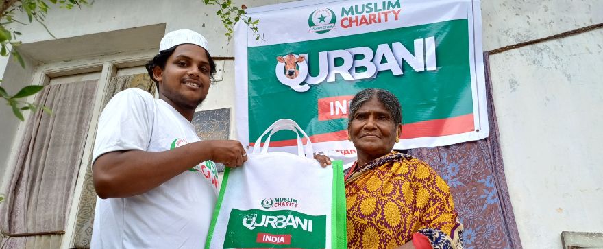 A man in a white cap and shirt hands a Qurbani food package to an elderly woman in a yellow-patterned sari, standing in front of a sign that reads "Qu