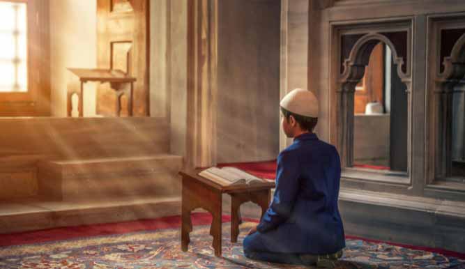 Young boy praying in a mosque, reading Quran on a wooden stand.